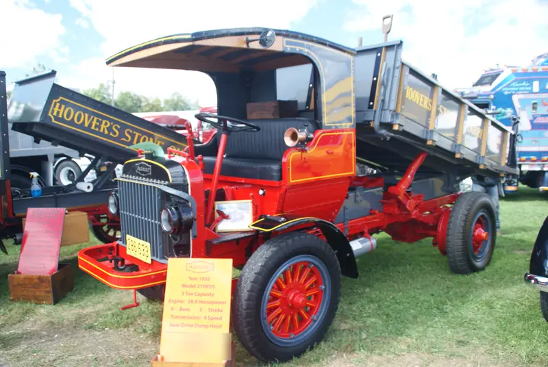 A restored red and black 1926 Autocar dump truck with an open cab, spoked red wheels and its wooden-sided dump body raised, parked on the grass at a truck show