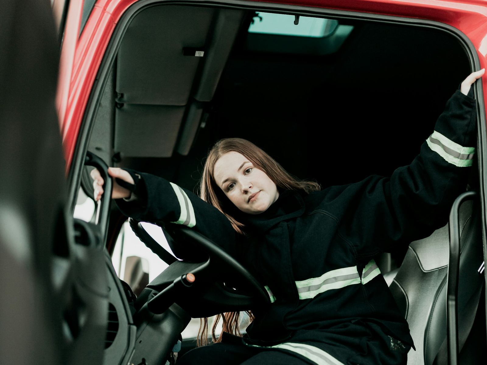 A driver in a reflective work jacket sits in the cab of a red truck, looking toward the camera