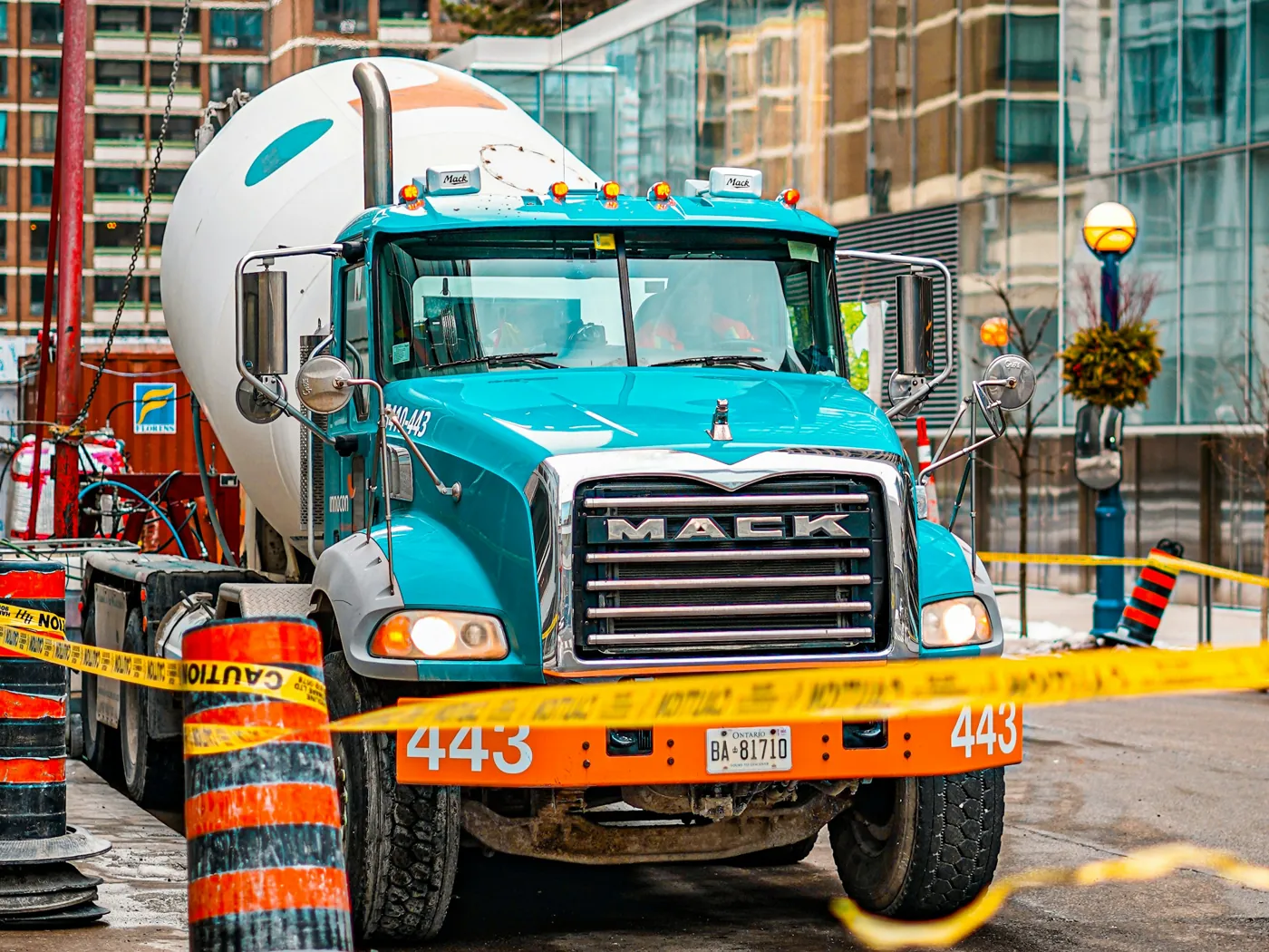A teal Mack concrete mixer truck at a downtown construction site behind caution tape and traffic barrels, with high-rise buildings close behind it