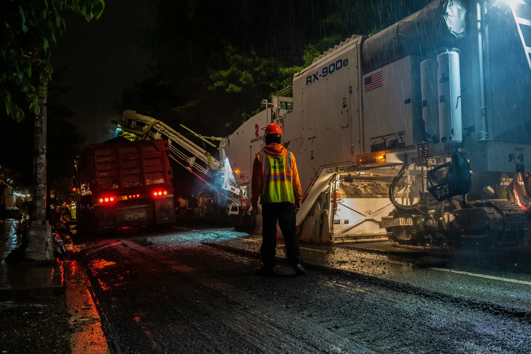 On a rainy night, a dump truck is loaded by a road milling machine’s conveyor while a worker in an orange hard hat and high-visibility vest stands in the street