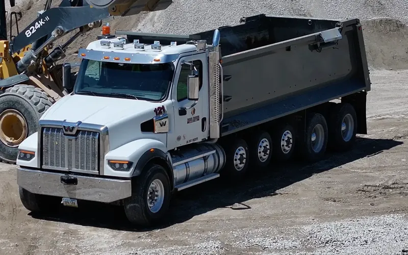 A wheel loader dumps aggregate into a white Western Star quint dump truck with a steer axle and five axles behind the cab at a quarry stockpile