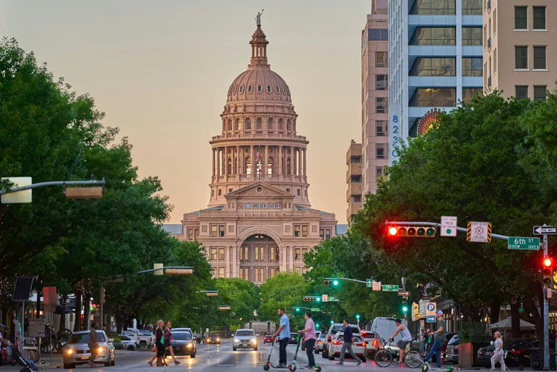 The Texas State Capitol in Austin at dusk, seen down Congress Avenue past traffic lights, cars and people crossing the street