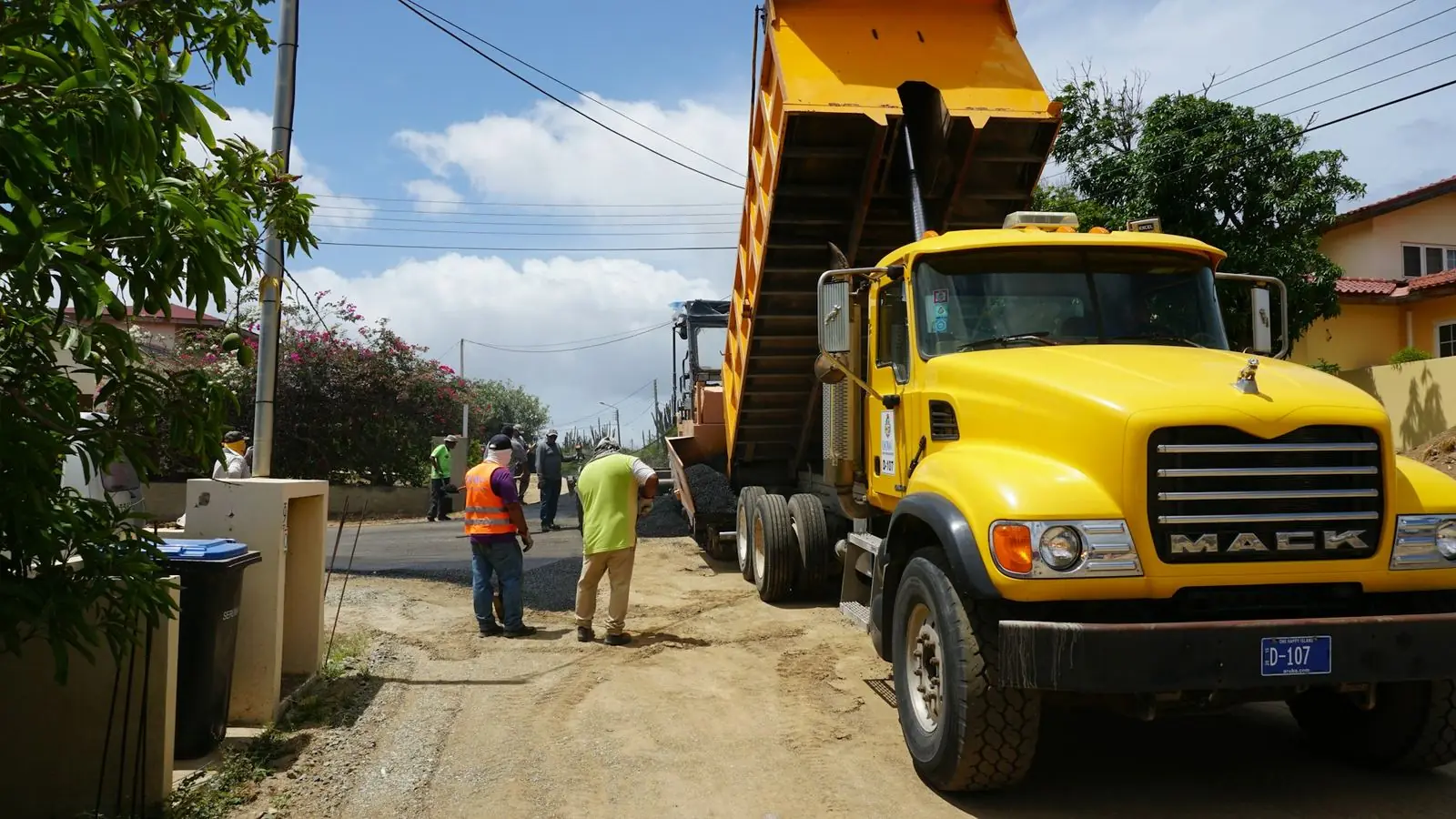 A yellow Mack dump truck with its body raised high below overhead utility lines on a narrow street while crew members work nearby
