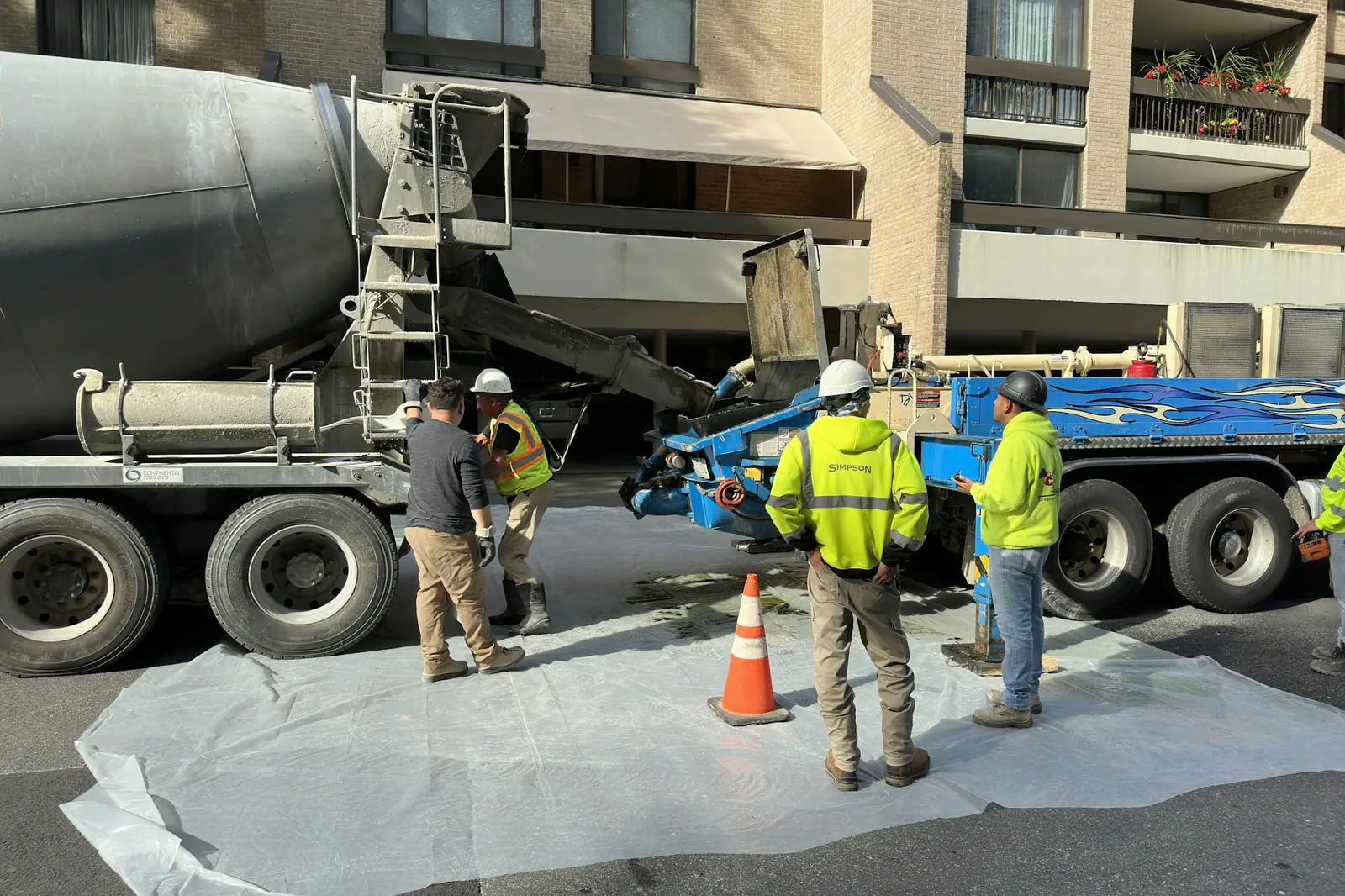 A ready-mix crew in hard hats and high-visibility vests and shirts sets up a pour beside a concrete mixer truck and a line pump on a city street