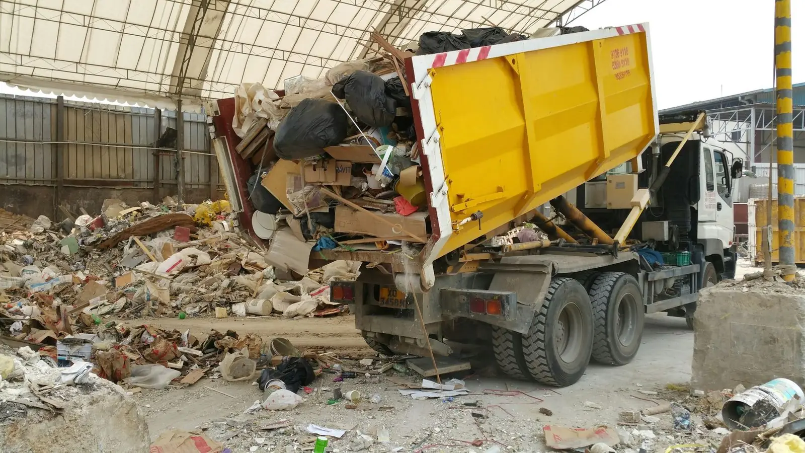 A yellow roll-off container tips a load of construction debris at a covered transfer station