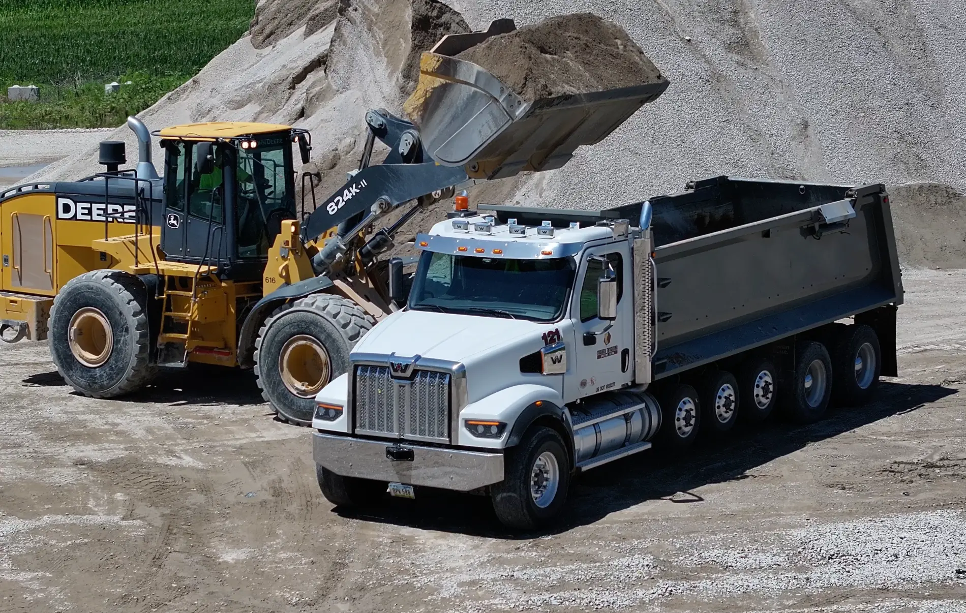 A wheel loader dumps sand into a white multi-axle dump truck at an aggregate yard