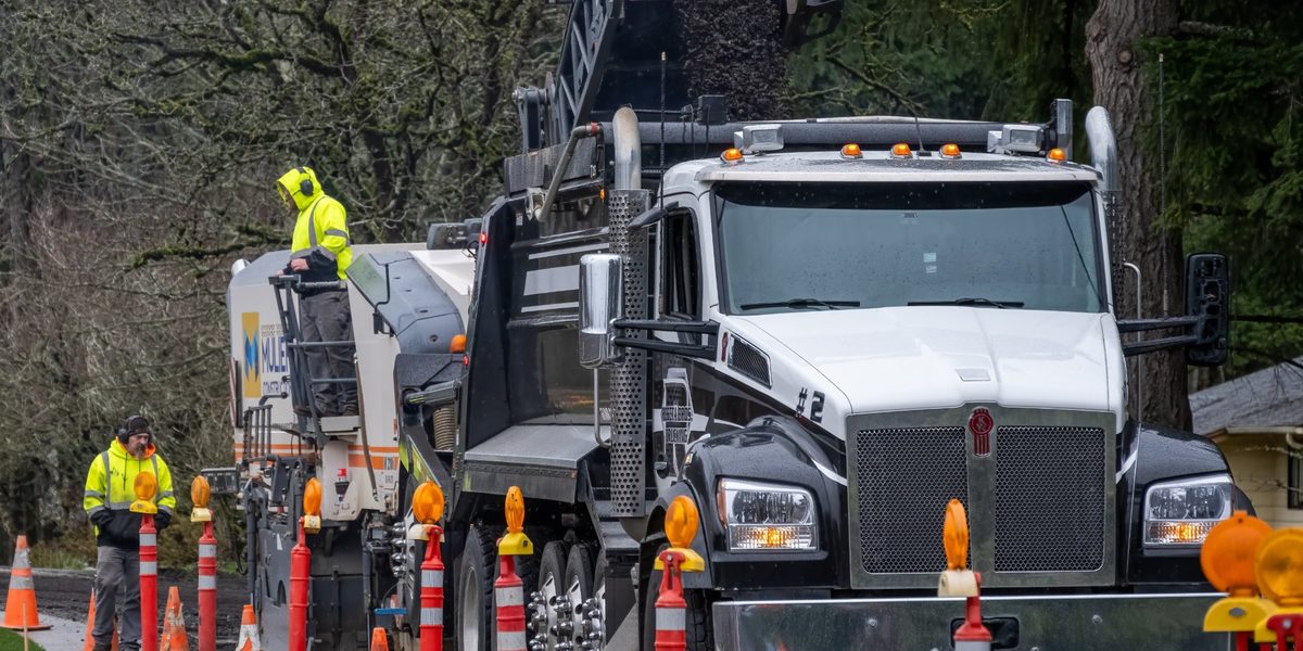 Dump truck being loaded with asphalt millings in a roadside work zone, with crew members in high-visibility gear