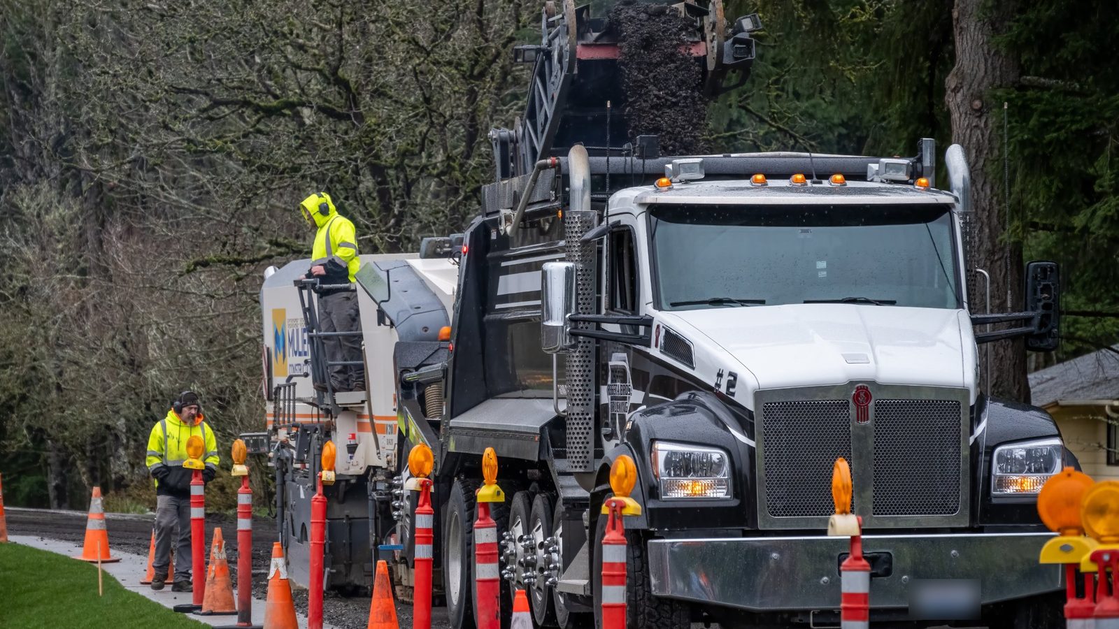 Dump truck being loaded with asphalt millings in a roadside work zone, with crew members in high-visibility gear