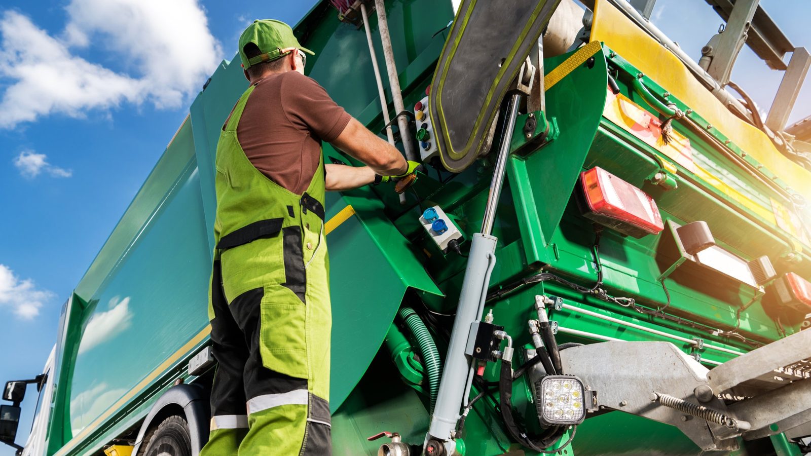 A refuse worker in high-visibility overalls works the packer controls at the back of a green rear-loader garbage truck