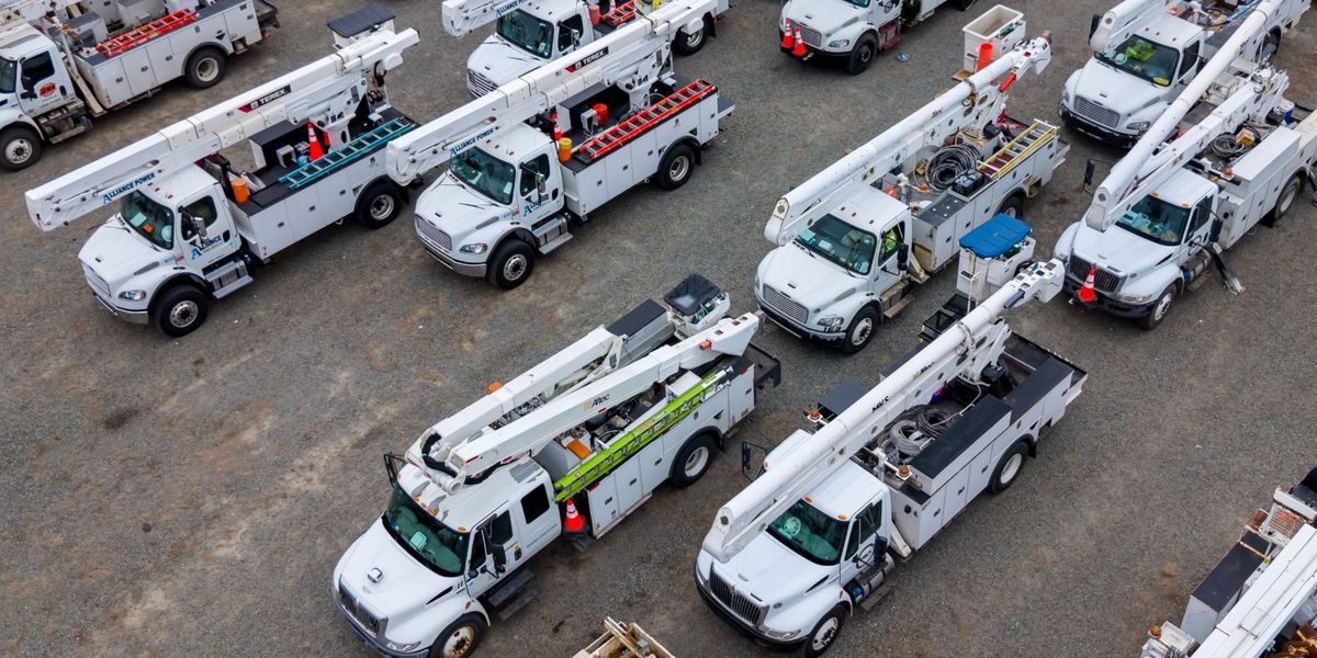 Rows of white bucket trucks and digger derricks, with orange tree-trimming trucks behind, staged in a gravel yard for storm restoration work