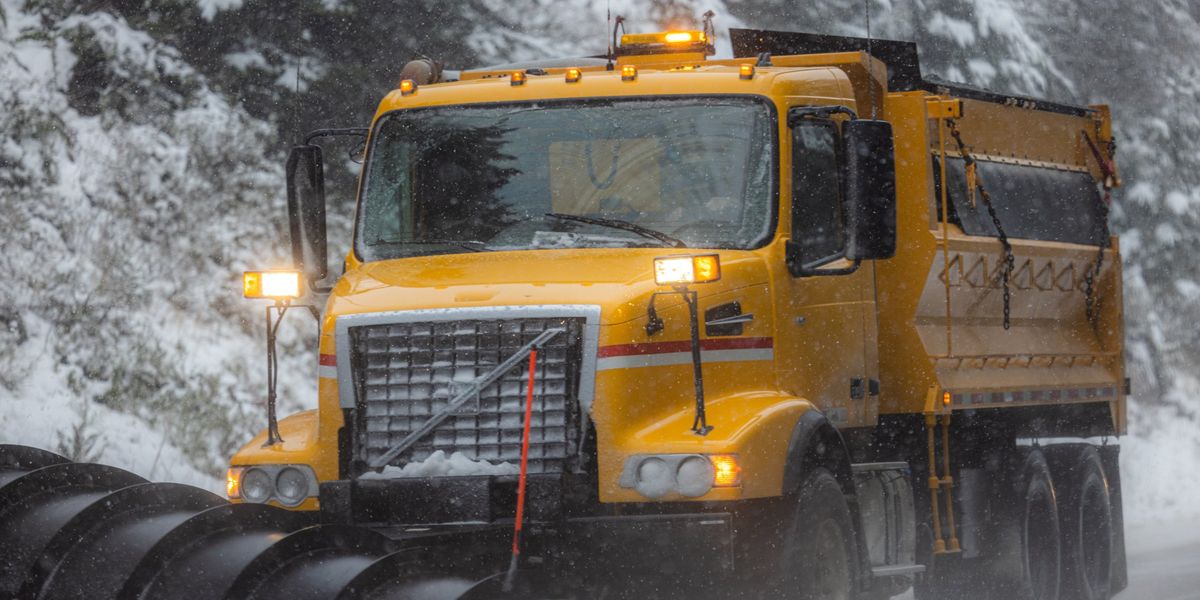 A yellow plow truck with a wing plow and salt spreader clears a mountain road during a snowstorm, amber lights flashing