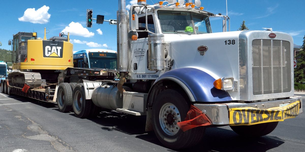 A Peterbilt tractor hauls a CAT excavator on a lowboy trailer with an oversize load banner through Flagstaff, Arizona