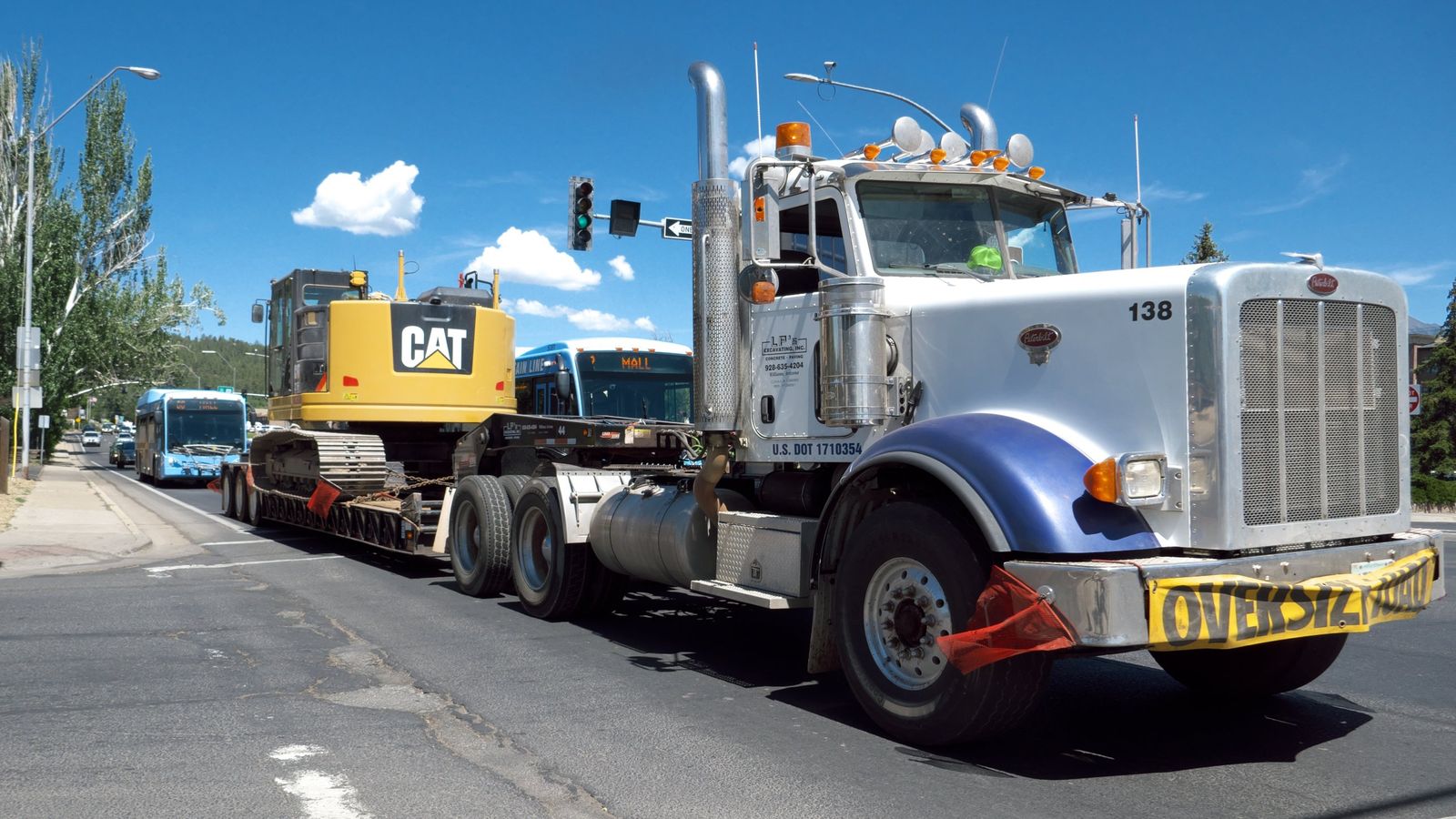 A Peterbilt tractor hauls a CAT excavator on a lowboy trailer with an oversize load banner through Flagstaff, Arizona
