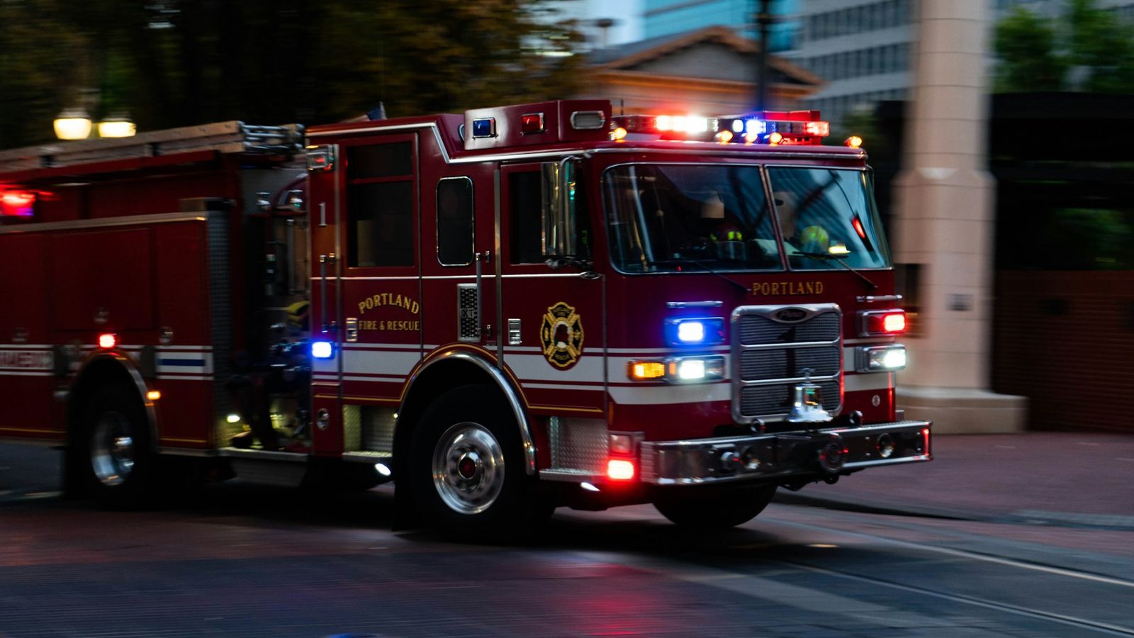 A Portland Fire & Rescue engine runs with red and blue lights flashing on a city street at dusk