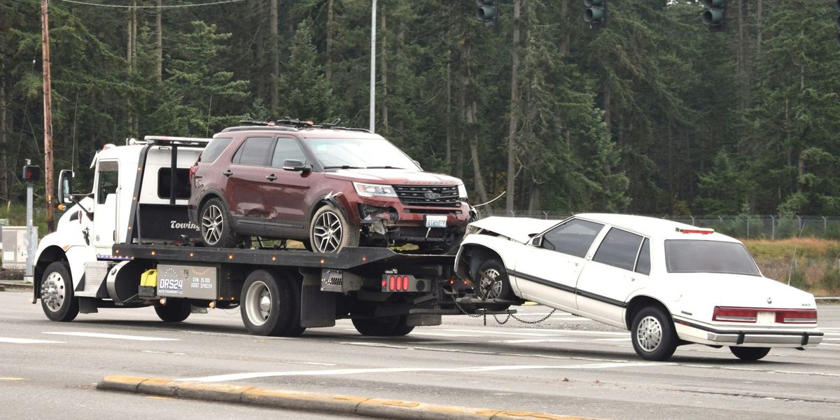 A rollback tow truck carries a damaged SUV on its deck while towing a crashed sedan behind it