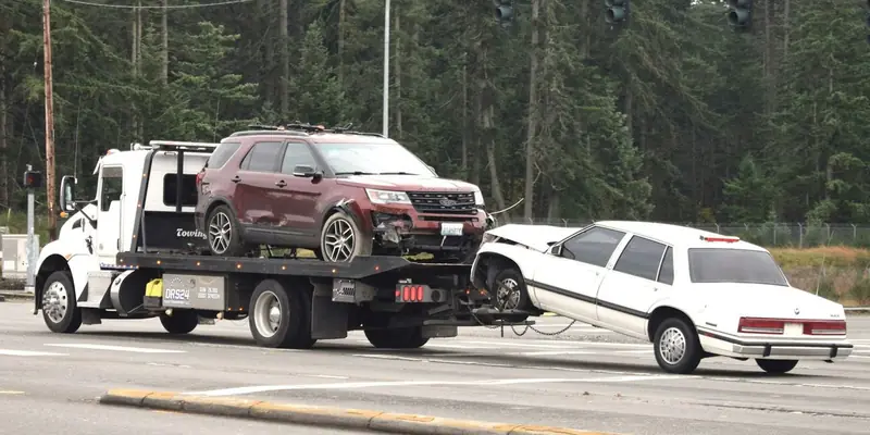 A rollback tow truck carries a damaged SUV on its deck while towing a crashed sedan behind it