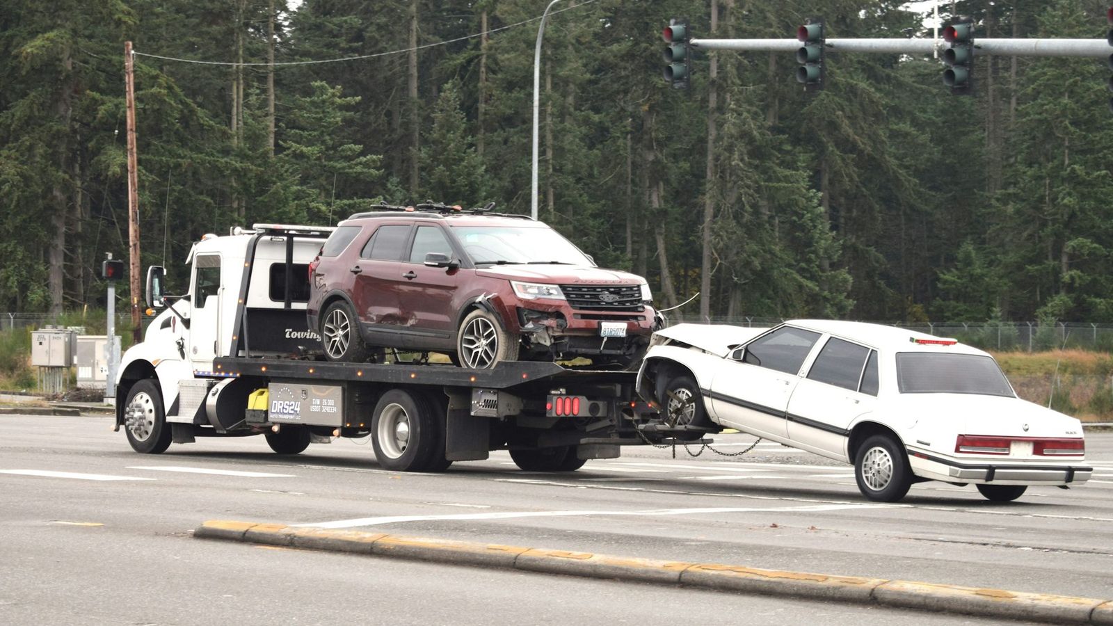 A rollback tow truck carries a damaged SUV on its deck while towing a crashed sedan behind it
