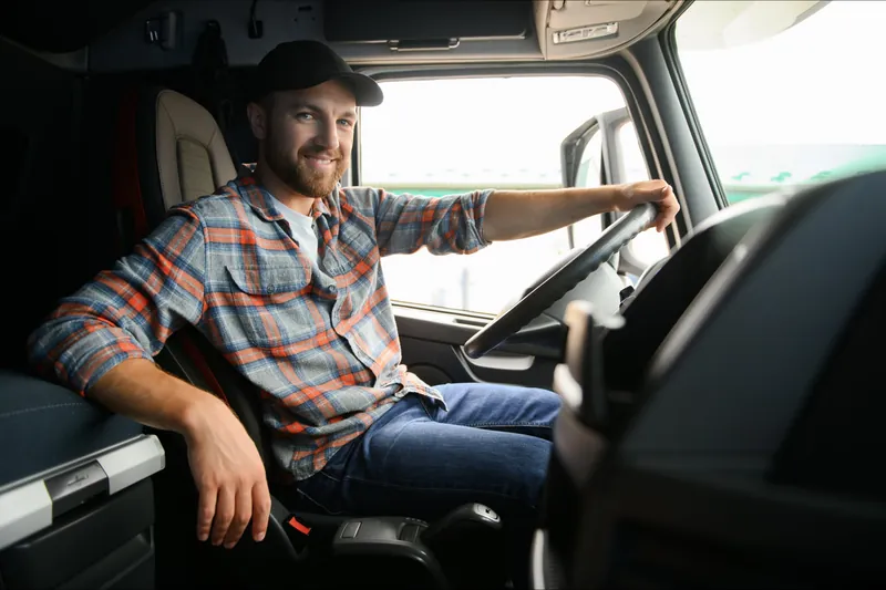 A smiling bearded driver in a plaid shirt and black cap sits behind the wheel in a truck cab